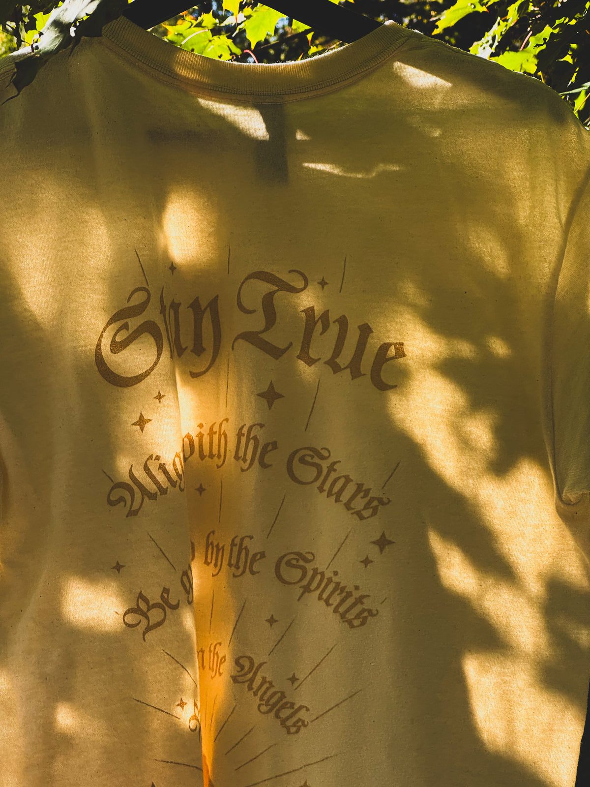 A beige t-shirt with a dark brown gothic-style graphic reading 'Stay True' under a sunburst design, photographed outdoors under dappled tree shadows.