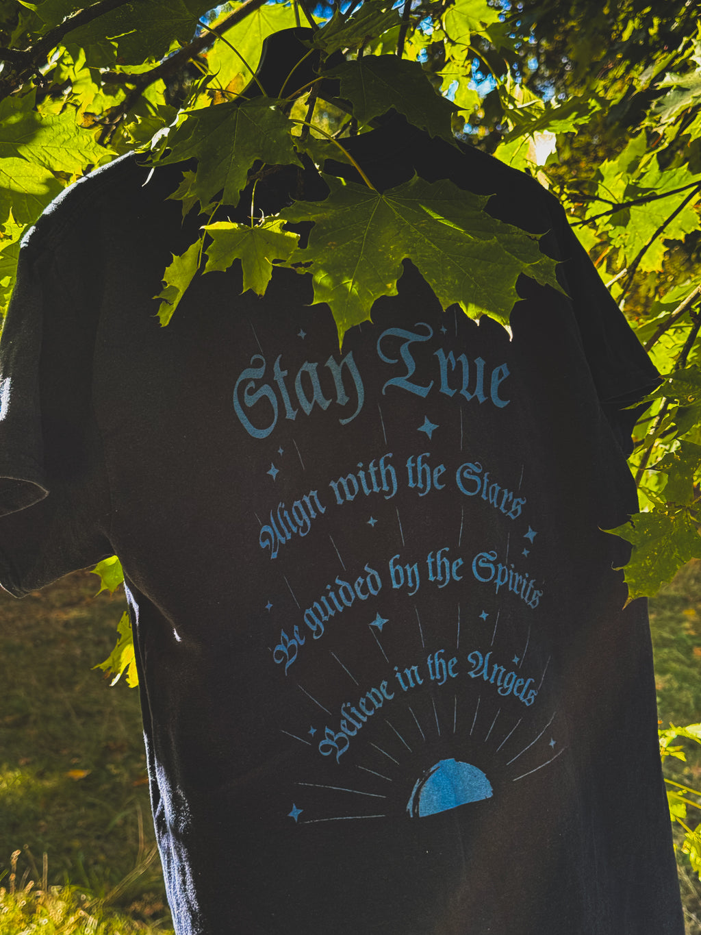 A navy graphic t-shirt hanging outdoors under green maple leaves, featuring a light blue gothic-style text design on the back.